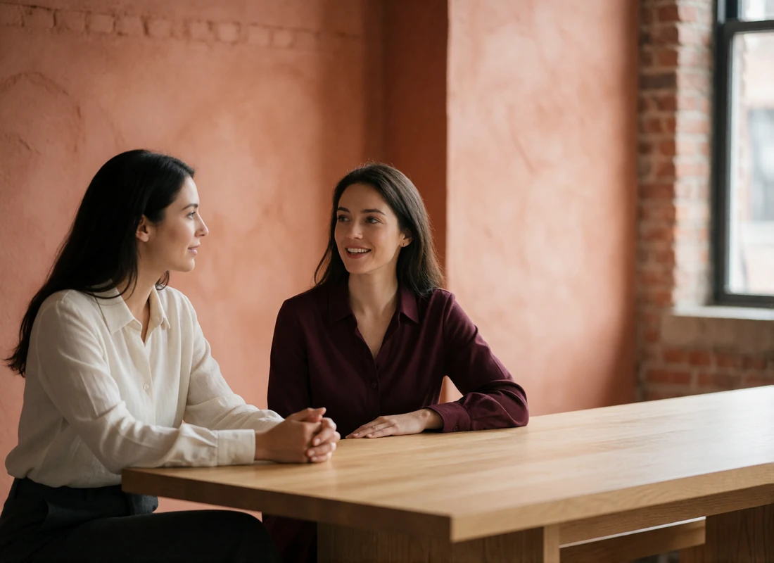 Two women in smart-casual clothing in conversation across a light oak table in a bright upscale workspace